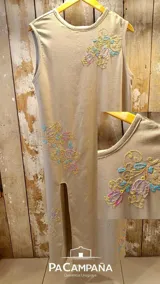 Vestido midi sin mangas de lino de algodón color beige, con cuello redondo y tajo lateral. Presenta bordados artesanales a mano de flores en tonos pastel en el pecho y en la parte inferior de la falda.