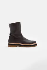 Bota de caña media confeccionada en cuero vacuno texturizado color marrón oscuro. Presenta un diseño minimalista con costuras reforzadas, tirador trasero y una suela de plataforma plana con capas en tonos beige, negro y suela.