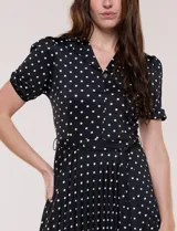 Vestido midi negro con estampado de lunares blancos, de la marca Sharagano. Presenta cuello con solapa, escote en V, cierre frontal con botones y mangas cortas. Tiene un corte en la cintura con lazo ajustable y falda plisada.