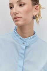 Camisa de algodón color celeste, con cuello mao y puños con detalle de volados. Presenta cierre frontal oculto con botones y un diseño de corte clásico.