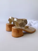 Sandalias de cuero color dorado, con tira ancha sobre los dedos, pulsera al tobillo con hebilla metálica y taco de madera.