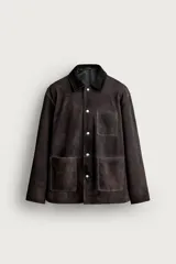 Cazadora de corte relaxed fit confeccionada en piel de ante de color marrón oscuro, con efecto lavado. Cuenta con cuello de solapa, manga larga, bolsillos frontales de parche y cierre frontal con botones.