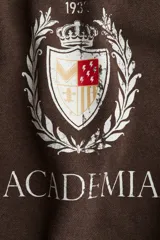 Buzo de tejido de punto color marrón oscuro con efecto lavado, cuello redondo y hombros caídos. Presenta un estampado frontal con el texto "MILANO" en tipografía universitaria, un escudo con la inscripción "193" y la palabra "ACADEMIA" debajo.