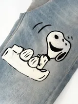 Jean de corte wide leg en denim azul claro con lavado desgastado. Presenta una ilustración pintada a mano de Snoopy en la pierna derecha, acompañada por la palabra 'Real', una corona y una estrella. Incluye etiqueta de cuero en la cintura y en el ruedo.