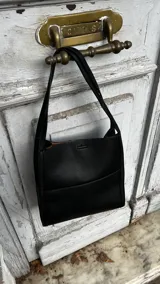 Cartera de mano tipo tote en color negro, confeccionada en cuero con textura granulada. Presenta un diseño minimalista con doble asa superior y un compartimento interior en tono contrastante.