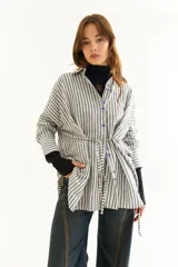 Camisa oversize de corte holgado con estampado de rayas verticales. Presenta cuello camisero, cierre frontal con botones a contraste y cordones integrados en la cintura para ajustar la silueta. Posee mangas largas con puños rectos.