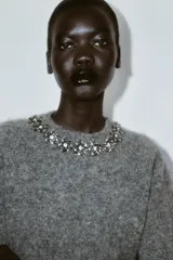 Jersey de punto de manga corta en color gris jaspeado, con cuello redondo decorado con abalorios joya y cierre de botón en la espalda.
