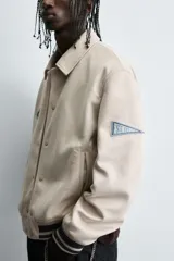 Cazadora estilo varsity de tejido efecto ante en color beige. Presenta cuello solapa, cierre frontal con botones a presión y puños y ruedo con acabado en rib a rayas negras. Incluye parches bordados decorativos en el pecho y la manga, además de bolsillos laterales de doble vivo.