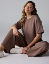 Conjunto comfy de buzo y pantalón wide leg color marrón, confeccionado en mezcla de poliéster, viscosa y elastano. El buzo es de silueta holgada, con escote redondo y mangas cortas de hombro caído. El pantalón es de tiro alto con pierna ancha y recta.