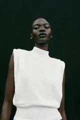 Top de punto blanco sin mangas con cuello subido, hombreras marcadas y espalda descubierta con cierre de botones.