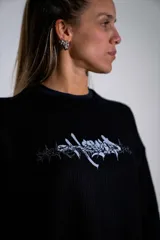 Buzo de tejido de punto fino en color negro, con diseño de cuello redondo y corte holgado. Presenta un estampado gráfico estilo gótico en el centro del pecho.