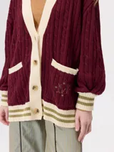 Cardigan de punto trenzado en color negro con detalles en color crema en los bolsillos, puños y ruedo. Presenta un diseño oversize, cierre frontal con botones y bordado decorativo en uno de los bolsillos delanteros.