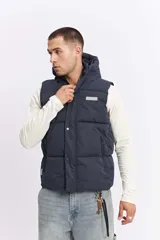Chaleco puffer acolchado color azul marino, con capucha fija, cierre frontal con solapa de botones a presión y bolsillos laterales con cierre. Presenta un parche con el logo de la marca en el pecho.
