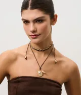 Collar de diseño multicapa que combina una cadena corta de eslabones redondos dorados con tiras de cordón marrón oscuro ajustables con terminales metálicas y dijes colgantes que incluyen una medalla texturizada, un corazón y una pieza en forma de colmillo.