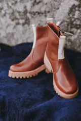 Botas de caña baja color marrón de cuero, con suela track ancha en tono tostado y detalle de franja elástica vertical en color blanco en la parte frontal. Cuentan con cierre lateral y tirador en la parte trasera.
