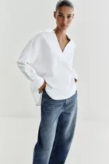 Blusa blanca confeccionada en lino, con cuello polo, escote en V y mangas largas amplias. Presenta un diseño holgado y bajo asimétrico.