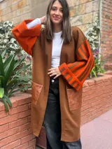 Saco largo de lana merino en color marrón, con diseño abierto sin botones. Presenta mangas anchas con estampado de rayas verticales en tonos naranja y marrón, y un bolsillo frontal aplicado en material símil cuero con textura.