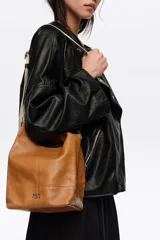 Bolso de mano Bimba y Lola modelo Paper mediano, confeccionado en cuero color tostado con estructura blanda, asas superiores y correa de hombro ajustable en color beige. Presenta costuras visibles, logo metálico de la marca en la parte inferior y cierre principal.