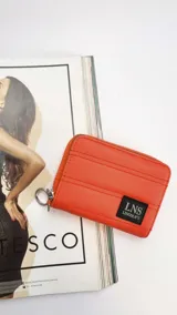 Billetera corta de diseño capitoneado con cierre perimetral y solapa frontal. Cuenta con compartimentos internos para tarjetas y billetes, e incluye una correa de mano desmontable.