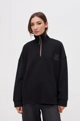 Buzo oversize de felpa con friza, color negro, con cuello alto y cierre frontal parcial metálico. Presenta bolsillos laterales, tajos en los costados y un diseño con espalda más larga. Incluye un pequeño logo bordado en el pecho.