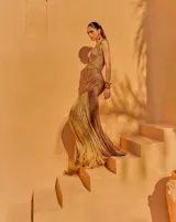 Vestido largo de tejido crochet en color bronce, con escote en V profundo, diseño de red y detalles trenzados. Presenta una silueta ajustada y un acabado con flecos largos en el ruedo.