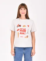 Remera de algodón color beige con estampado frontal temático de París que incluye texto y diversos iconos como una torre Eiffel, una scooter, un croissant y un perro.