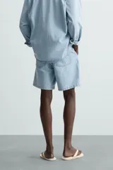 Shorts de denim de algodón, color celeste lavado, de corte regular fit y largo hasta la rodilla. Presenta cierre frontal con cremallera y botón, bolsillos delanteros y bolsillos traseros de plastrón. El borde inferior tiene un acabado deshilachado.