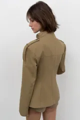 Blazer entallada color beige con cuello subido, manga larga con hombreras y trabillas en los hombros. Presenta bolsillos de plastrón en el delantero, detalle de solapas en el pecho y cierre frontal con botones a presión.