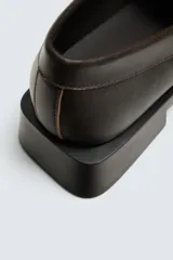 Mocasín de vestir color marrón oscuro, con empeine liso y detalle de antifaz. Presenta horma cuadrada y suela con ligero tacón geométrico a contraste.
