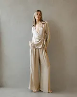 Conjunto de dos piezas en tono arena con textura plisada y acabado satinado. La parte superior es una blusa cruzada de manga larga con escote en V profundo y detalle de hebilla lateral. El pantalón es de corte wide leg con caída fluida.