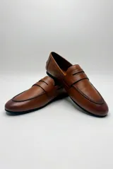 Mocasín de cuero color café, diseño clásico de estilo penny loafer con suela plana y costuras visibles.