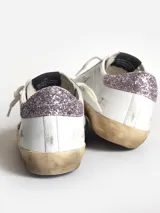 Zapatillas Golden Goose modelo Super-Star, de cuero blanco con estrella de cuero color cuero, refuerzo del talón con purpurina rosa, lengüeta de ante gris hielo y cordones color crema.
