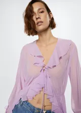 Vestido largo de tejido fluido y semitransparente color lila, con cuello en pico, mangas largas con volantes y cierre frontal con lazo. Presenta volantes decorativos a lo largo de la abertura frontal y fruncido elástico en la cintura.