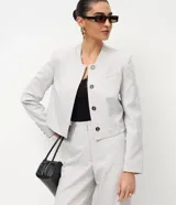 Chaqueta de sastre estructurada color gris claro, con cuello redondo tipo rectilínea, cierre frontal con botones y mangas largas con botones en los puños. Presenta un corte levemente corto y diseño minimalista.