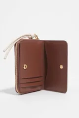 Billetera Bimba y Lola color marrón, de estructura rígida en piel vacuna, con cierre de cremallera y tirador de cordón en color crudo. Incluye logo de la marca estampado en dorado en el frente.