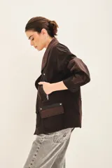 Campera de corte holgado en color marrón oscuro, con cierre frontal mediante broches y jareta interna para ajuste en la cintura. Presenta un diseño con cuello solapa y detalles de costuras visibles.