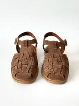 Sandalias estilo cangrejera color marrón, confeccionadas en cuero con diseño trenzado en la parte delantera, tira ajustable con hebilla metálica en el tobillo y suela plana.