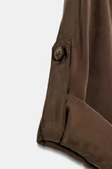 Vestido midi de corte fluido en color marrón, con cuello solapa, escote en pico y cierre frontal de botones. Presenta mangas cortas con dobladillo, bolsillos delanteros y un cinturón trenzado con hebilla redonda de madera.