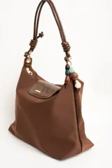 Cartera tipo shopper color marrón, con solapa frontal decorativa en tono más oscuro y tiras con nudos. Incluye herrajes dorados y un charm colgante con cuentas de madera.