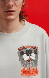 Remera de algodón premium color gris claro, corte oversize, con cuello redondo y manga corta. Presenta un estampado frontal pequeño con dos personajes de Disney (Mickey y Minnie) en llamas con el texto "THE FLAME OF LOVE".
