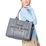 Cartera tipo tote de cuero granulado en color gris, con diseño estructurado y asas superiores. Presenta el logo estampado en el frente con la inscripción The Tote Bag Marc Jacobs.