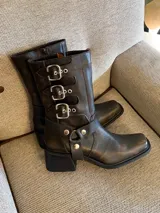 Bota de cuero vacuno color marrón oscuro, de caña media, con punta cuadrada y taco bajo. Presenta correas con hebillas metálicas plateadas y estribo desmontable.