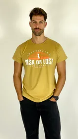 Remera de manga corta color amarillo mostaza con cuello redondo. Presenta un estampado frontal con el texto 'Risk Or Lost' y un gráfico de un ave con sombrero sobre un sol poniente.