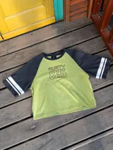 Remera bicolor Rusty con cuerpo verde lima y mangas ranglán grises con dos rayas blancas en los puños. Estampado frontal con el logo de la marca y el número 85.
