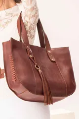Bolso tipo shopper de cuero vacuno color tabaco, con diseño amplio y estructurado. Presenta detalles de trenzado artesanal en los laterales, asas largas para hombro y un colgante decorativo con flecos de cuero y herrajes metálicos.