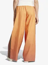 Pantalón deportivo Adidas de corte holgado con diseño degradado en tonos naranja y marrón. Presenta las tres rayas negras características de la marca en los laterales y logo bordado en la parte superior.
