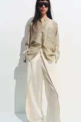 Camisa de cuello mao color beige, confeccionada en tejido satinado con brillo sutil, manga larga con puños anchos y un bolsillo de parche en el pecho con detalles bordados tonales. Cierre frontal con botones ocultos bajo solapa.