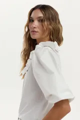Blusa blanca con cuerpo de jersey y mangas de tela plana abullonadas. Cuenta con volados en los hombros decorados con bordados en contraste.