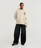 Hoodie color beige claro, corte boxy, con capucha ajustable y bolsillo canguro. Presenta un gran estampado gráfico en la espalda con un dragón oriental, texto en inglés y caracteres japoneses, y la palabra "BRAVE" en la parte superior.
