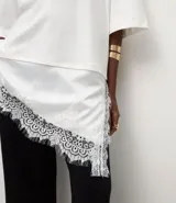Remera de algodón corte oversized, color blanco, con cuello redondo, mangas cortas amplias y ruedo asimétrico con detalle de encaje aplicado.